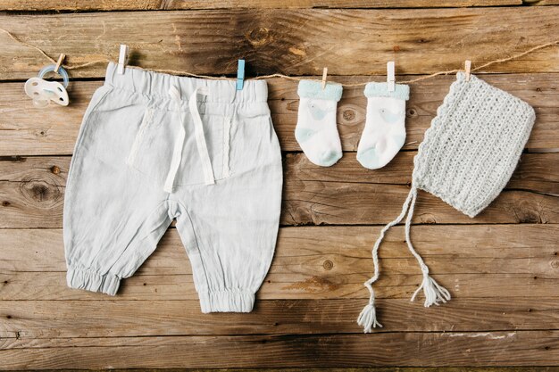Calça do bebê; meias; headwear e chupeta pendurado no varal com prendedores de roupa contra a parede de madeira
