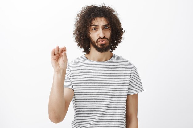 Cale a boca, por favor. Retrato de modelo masculino bonito incomodado e irritado com barba e cabelo encaracolado, mostrando gesto de silêncio com a palma da mão