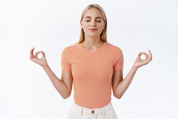 Calma e paciência Garota feminina caucasiana loira atraente em camiseta listrada feche os olhos e sorria satisfeito relaxando após um dia duro de trabalho meditando fazer exercícios de respiração fundo branco