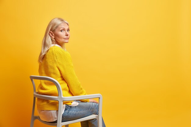 Calma, linda mulher de cinquenta anos sentada na cadeira sozinha, pensando na vida, usa suéter de malha amarela e jeans em branco cópia espaço
