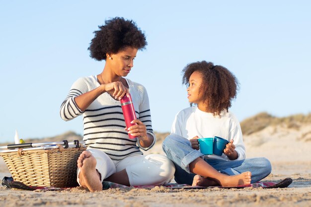 Calma mãe e filha afro-americanas no piquenique na praia. Mulheres em roupas casuais sentadas no cobertor, segurando garrafas térmicas e canecas. Família, relaxamento, conceito de natureza