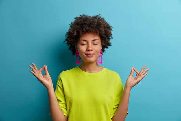 Calma relaxada mulher de pele escura fica em pose de lótus, sente-se aliviada, tenta se concentrar em uma atmosfera pacífica, usa roupas verdes casuais, isolada na parede azul. conceito de linguagem corporal