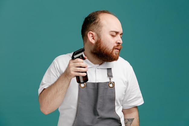 Calmo jovem barbeiro masculino vestindo camisa branca e avental de barbeiro virando de cabeça para o lado aplicando spray de cabelo em sua própria barba com os olhos fechados isolados no fundo azul