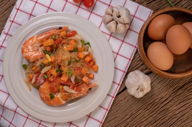 Camarão frito com macarrão de vidro em um prato branco colocado sobre um pano com ovos e alho.