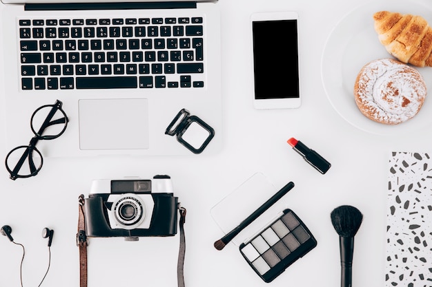 Câmera; computador portátil; celular; produto de cosméticos e pastelaria assada na mesa branca