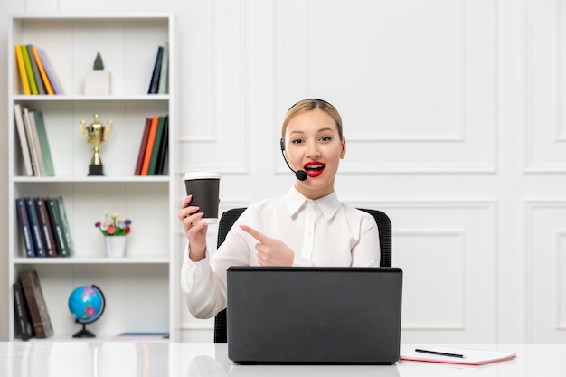 Camisa de escritório loira bonita de atendimento ao cliente com fone de ouvido e computador apontando para a xícara de café