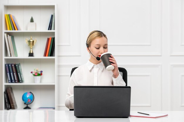 Camisa de escritório loira bonita de atendimento ao cliente com fone de ouvido e computador bebendo um café