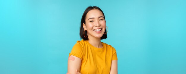 Campanha de vacina de covid19 Menina asiática feliz e saudável rindo após a vacinação de coronavírus bandaid no ombro vestindo camiseta amarela fundo azul