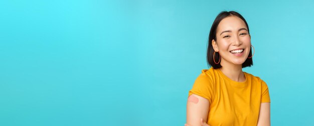 Campanha de vacina de covid19 Menina asiática feliz e saudável rindo após a vacinação de coronavírus bandaid no ombro vestindo camiseta amarela fundo azul