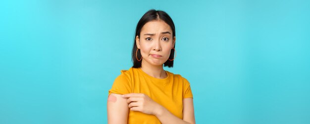 Campanha de vacina de covid19 Mulher asiática triste e decepcionada mostrando ombro com bandaid e careta chateada em pé sobre fundo azul