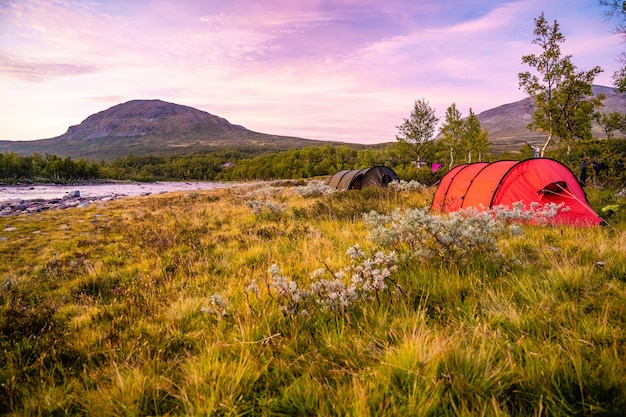 Campo com tendas rodeadas por colinas cobertas de verde sob um céu nublado durante o pôr do sol