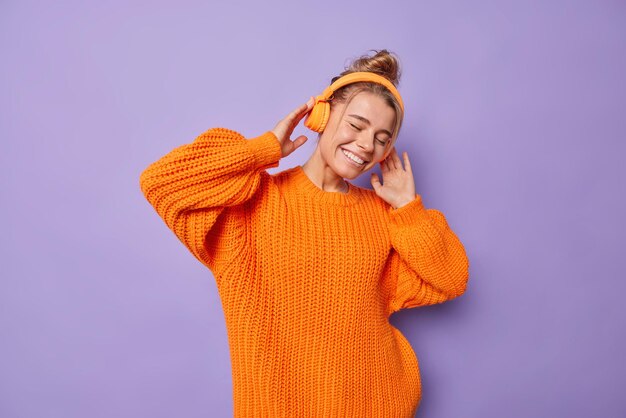 Cândida linda mulher Europeia se diverte mantém as mãos nos fones de ouvido, sendo feliz aprecia sua música favorita, vestida com danças de suéter laranja solto contra o fundo roxo. Ótima maneira de sentir prazer