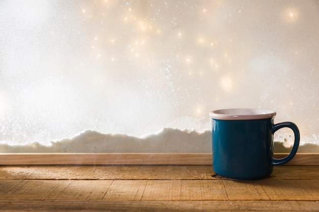 Caneca azul na mesa de madeira perto de banco de neve e luzes de fada