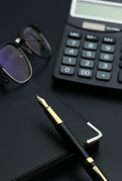 Caneta dourada, caderno, calculadora e óculos na mesa preta