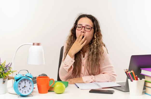 Cansada, jovem e bonita colegial de óculos, sentada na mesa com as ferramentas da escola, fazendo a lição de casa, colocando a mão na boca e bocejando com os olhos fechados, isolado no fundo branco