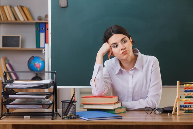 cansado de colocar a mão na bochecha jovem professora sentada na mesa com ferramentas escolares em sala de aula
