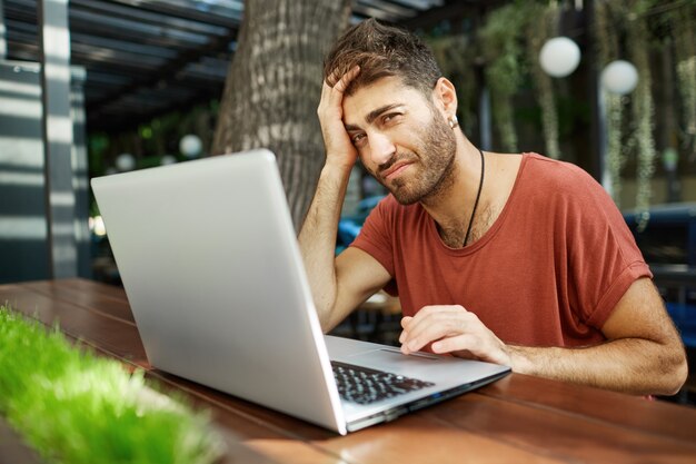 Cansado e barbudo freelancer bonito, programador ou estudante usando laptop enquanto está sentado em um café ao ar livre