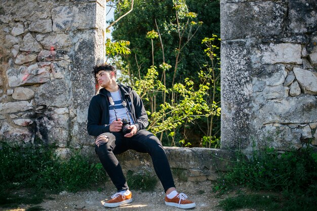 Cansado jovem fotógrafo sentado no velho muro de pedra com a câmera de exploração