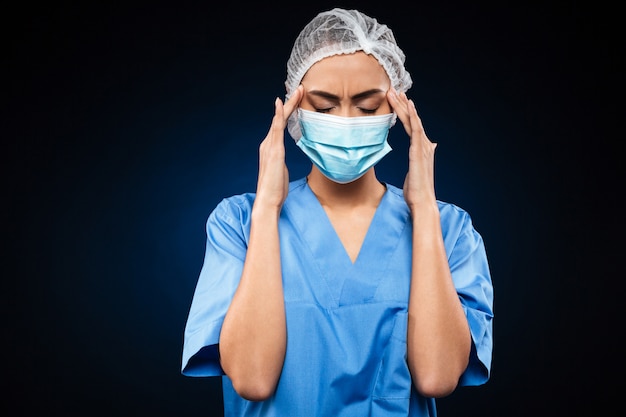 Cansado médico feminino em máscara médica e boné tem uma dor de cabeça isolada