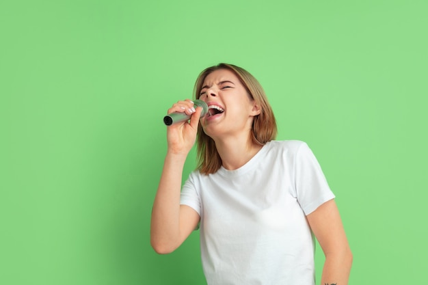 Cantando com alto-falante. Retrato de mulher jovem branca isolado na parede verde. Bela modelo feminino em camisa branca. Conceito de emoções humanas, expressão facial, juventude.