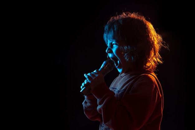 Cantando como celebridade, rockstar. retrato do menino caucasiano na parede escura em luz de néon. lindo modelo cacheado. conceito de emoções humanas, expressão facial, vendas, anúncio, música, hobby, sonho.