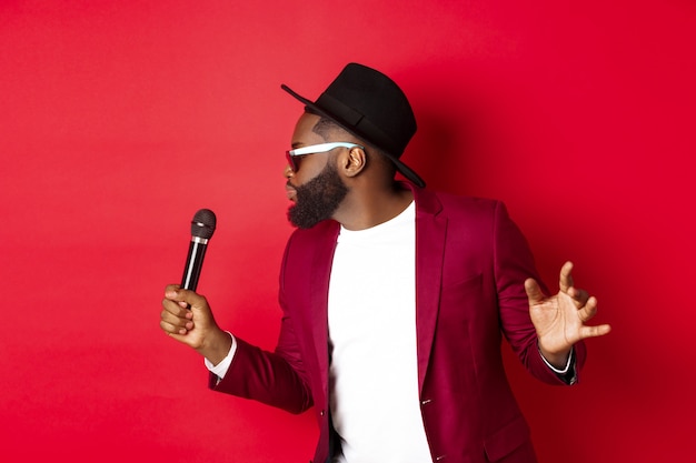 Foto grátis cantor negro apaixonado realizando contra um fundo vermelho, cantando no microfone, vestindo roupa de festa, em pé sobre um fundo vermelho.