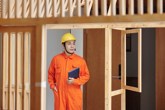 Capataz de macacão laranja e capacete de segurança segurando um tablet e andando pela casa em construção.