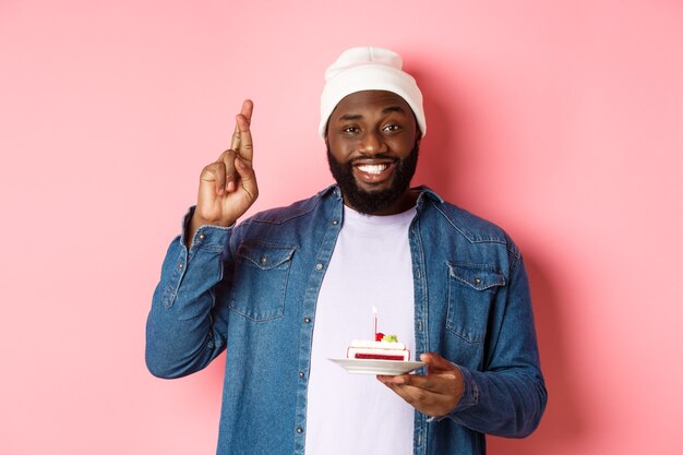 Cara afro-americano bonito comemorando aniversário, fazendo pedido com os dedos cruzados, segurando bolo de aniversário com vela, em pé contra um fundo rosa