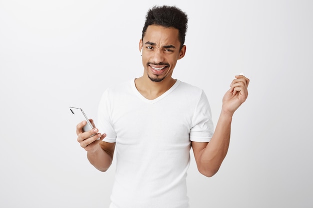 Cara afro-americano frustrado e descontente tirando o fone de ouvido e se encolhendo, segurando o celular