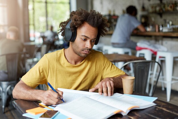Cara afro-americano hipster com livro e caderno ouvindo música em fones de ouvido e bebendo café enquanto está sentado em um restaurante aconchegante. Conceito de educação