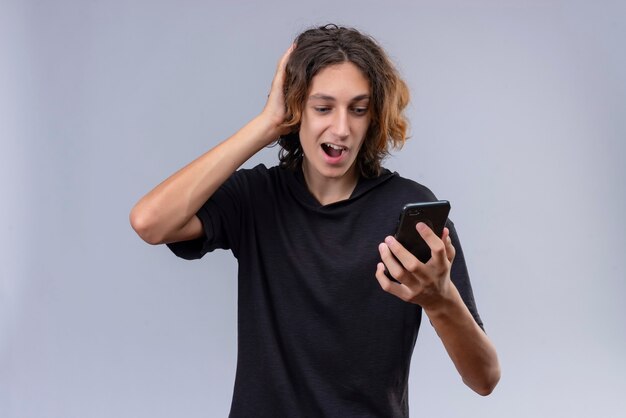 Cara alegre com cabelo comprido em uma camiseta preta segurando um telefone e agarrando a cabeça na parede branca