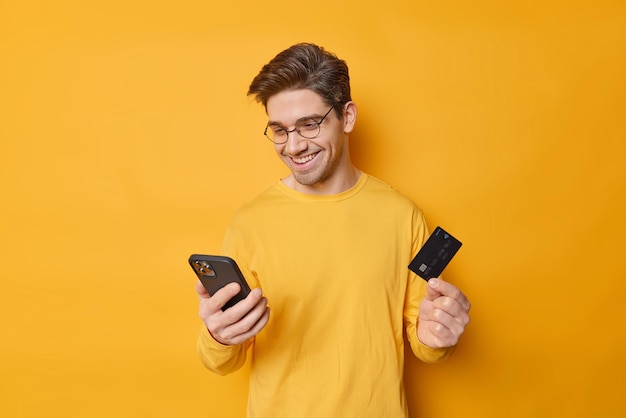 Cara alegre com cabelo escuro usa telefone celular e cartão bancário para fazer compras on-line, algo na loja on-line insere informações para pagamento seguro isolado sobre fundo amarelo