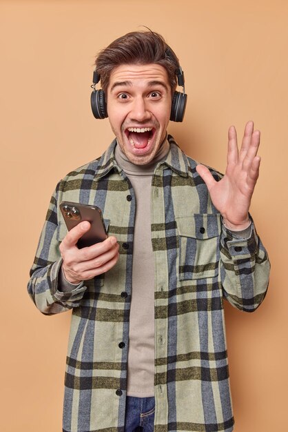 Cara alegre exclama em voz alta mantém a palma da mão levantada reage a notícias incríveis ouve música em fones de ouvido segura celular usa camisa quadriculada isolado ver fundo bege desfruta da lista de reprodução favorita