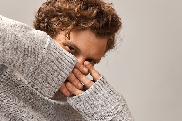 Cara atraente hipster com cabelo ruivo ondulado cruzando os dedos sobre o rosto com expressão facial penetrante, vestido com um pulôver cinza. copyspace para sua informação