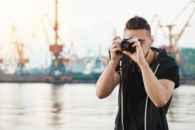 Cara atraente, trabalhando com a câmera. Jovem fotógrafo elegante olhando através da câmera durante a sessão de fotos com o modelo lindo, tirando fotos no porto perto da beira-mar, com foco no trabalho