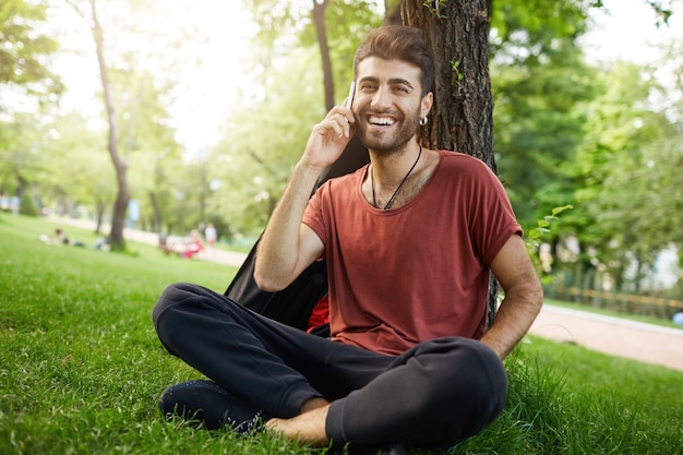 Cara barbudo bonito descansando no parque na grama, falando no celular e sorrindo feliz