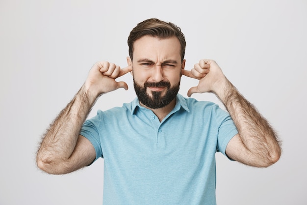 Cara barbudo perturbado fechando as orelhas e apertando os olhos irritado