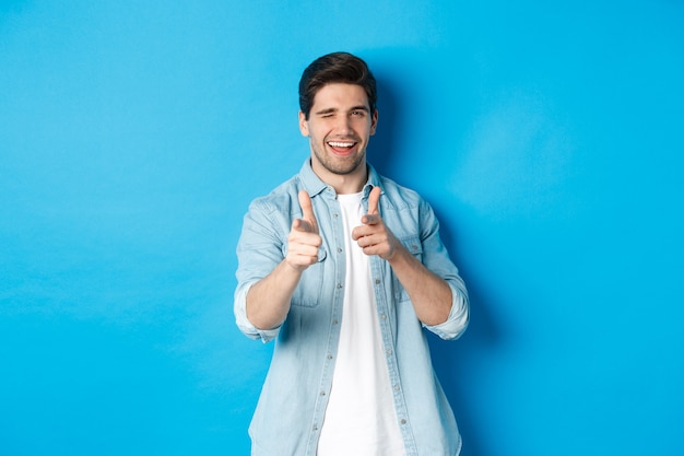 Foto grátis cara bonito atrevido apontando o dedo para você, piscando como um glamour, vestindo uma roupa casual contra um fundo azul