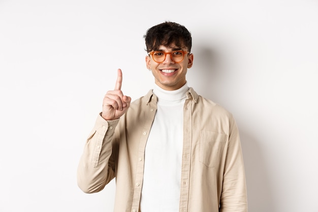 Cara bonito, caucasiano, de óculos, mostrando um dedo e sorrindo, em pé sobre um fundo branco