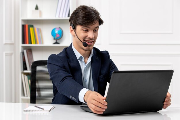 Cara bonito de atendimento ao cliente com fone de ouvido e laptop de terno feliz olhando para a tela