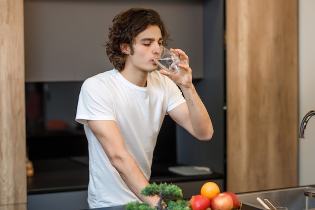 Cara bonito fica na cozinha moderna, bebendo um copo d'água de manhã