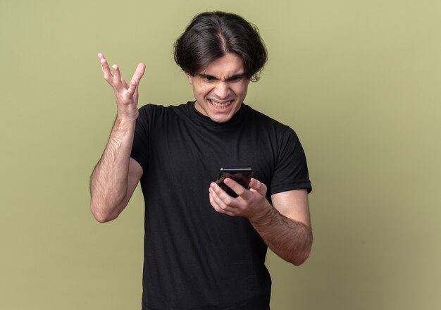 Cara bonito jovem bravo vestindo camiseta preta segurando e olhando para o telefone isolado na parede verde oliva