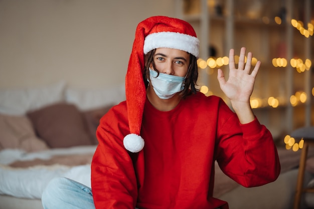 Cara caucasiana com máscara facial e chapéu de papai noel posando e olhando para a câmera em casa