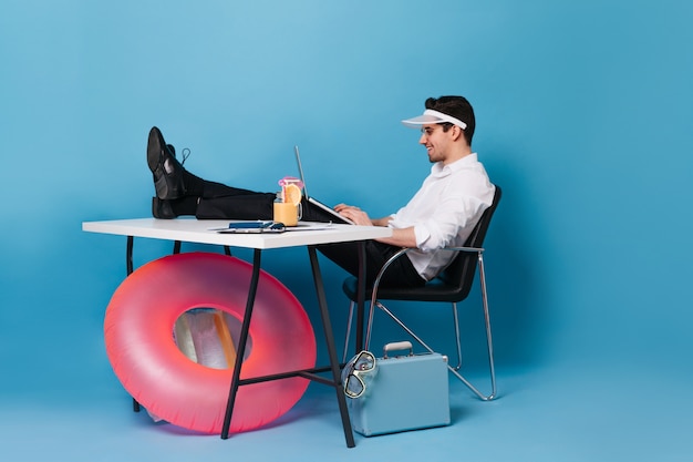 Cara de boné trabalha com laptop, sentado com as pernas jogadas na mesa. Retrato de homem contra o espaço da mala e do círculo inflável.