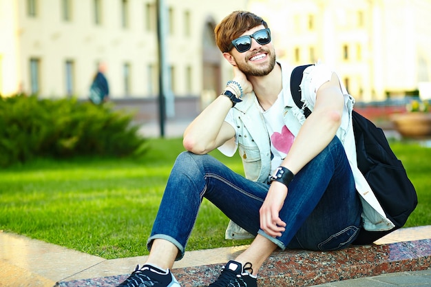 Cara de homem bonito hipster sorridente engraçado em pano elegante de verão na rua sentado na grama do parque