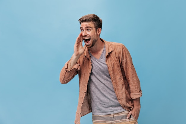 Foto grátis cara de olhos cinza positivos com barba legal em uma camisa da moda e calça listrada bege olhando para longe e gritando na parede azul