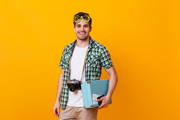 Cara de turista vestindo camisa xadrez e camiseta branca, olhando para a câmera. Retrato de homem com máscara de mergulho na cabeça, segurando a câmera retro e a mala de mão.