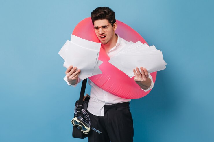 Foto grátis cara descontente de camisa branca, segurando uma pilha de papel de escritório. morena masculina posando com anel de borracha rosa contra o espaço azul.