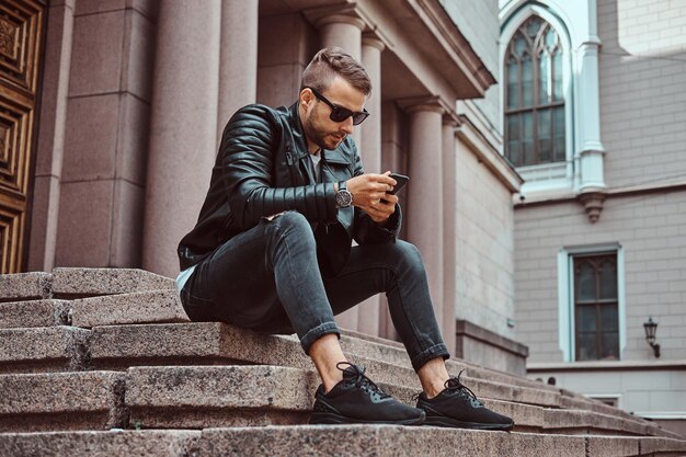 Cara elegante vestido com uma jaqueta preta e jeans usando um smartphone sentado nos degraus contra um prédio antigo na Europa.