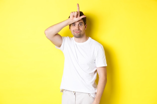 Cara estranho mostrando sinal de perdedor na testa, parecendo triste e sombrio, em pé com uma camiseta branca contra um fundo amarelo.